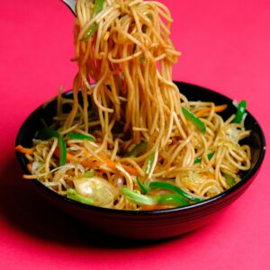 A serving of tasty Asian noodles with vegetables in a black bowl on vibrant pink background.
