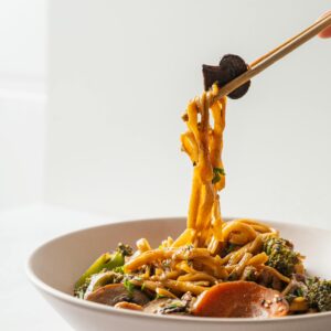 Close-up of chopsticks holding stir-fried noodles with vegetables in a bowl, perfect for food presentation.