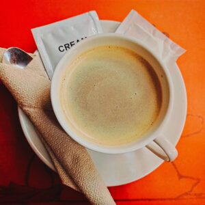 A steaming cup of coffee with creamer packets and a spoon on a vibrant orange background.